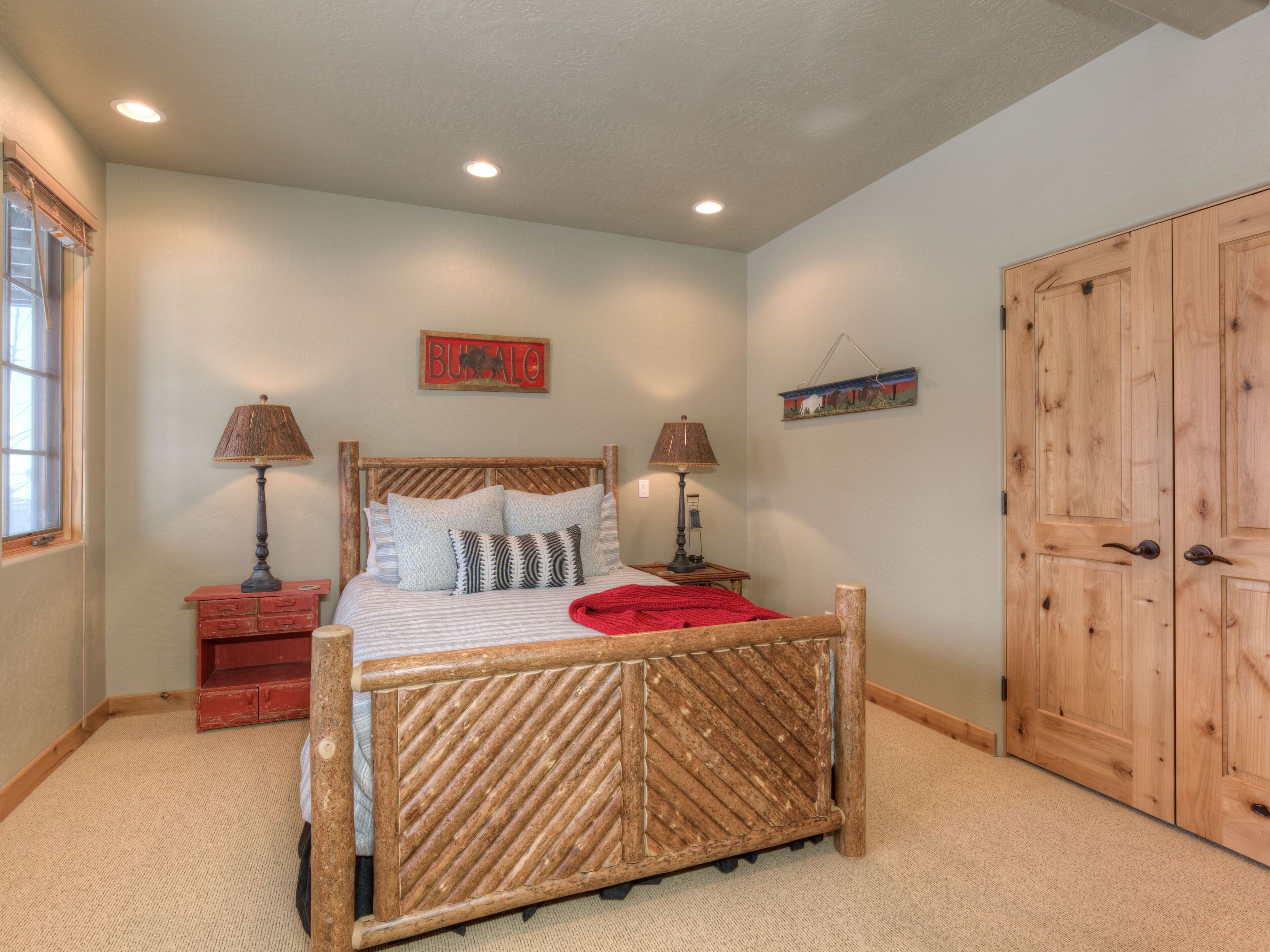 Une chambre à coucher avec un cadre de lit en bois, deux tables de chevet avec des lampes, une couverture rouge, un tableau accroché au mur et une armoire en bois.