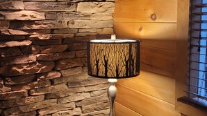 Una habitación con pared de piedra, paneles de madera, una mesa con libros, una lámpara con un diseño de sombra de árbol y una cabeza de venado montada.