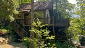 Une cabane à deux étages dotée d’une terrasse en bois et d’un escalier, entourée d’une forêt dense.