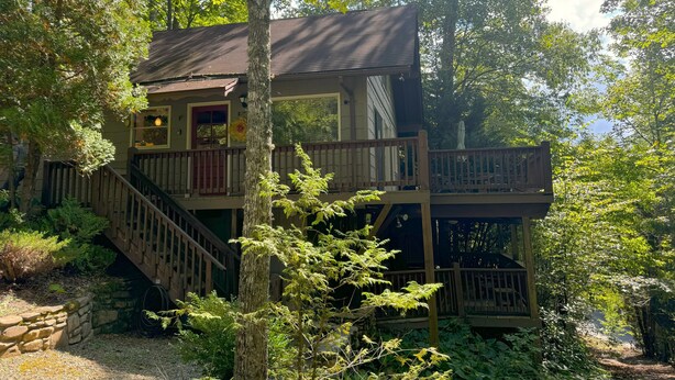 Una cabaña de dos pisos con terraza de madera y escaleras, rodeada de un bosque denso.