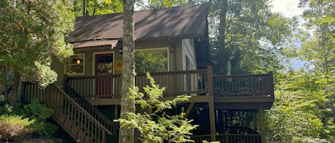 Une cabane à deux étages dotée d’une terrasse en bois et d’un escalier, entourée d’une forêt dense.