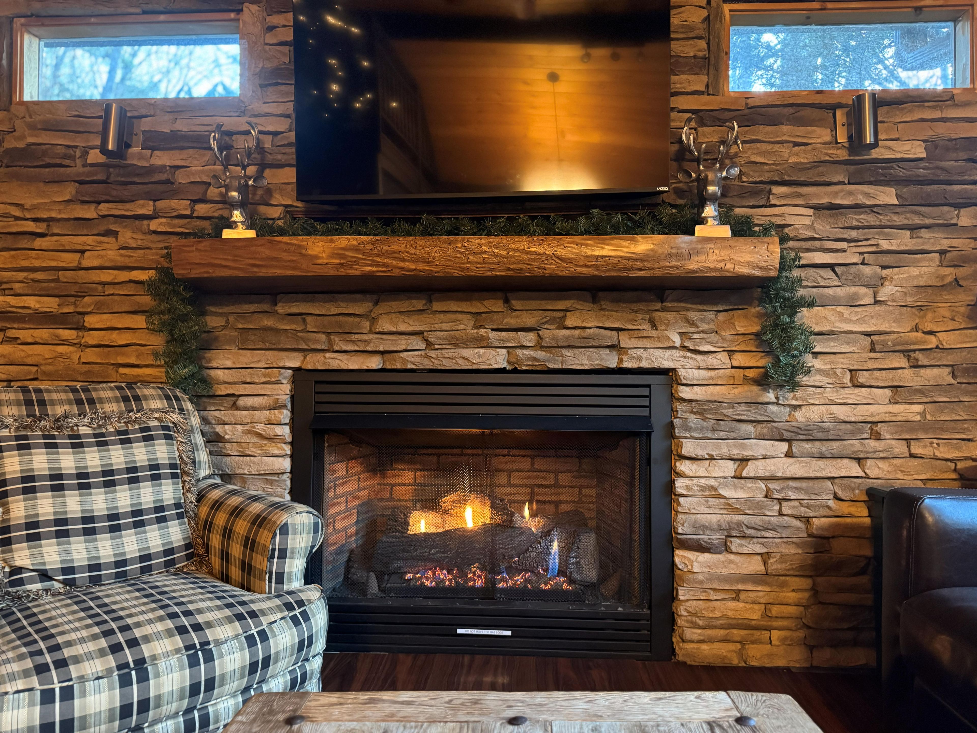 Cozy stone fireplace with TV, plaid chair and leather sofa—true cabin warmth.