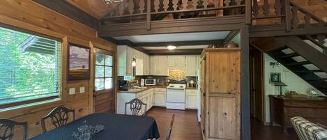 Interior rústico de una cabaña con comedor, cocina y un dormitorio en el altillo.