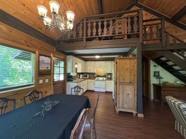 Interior rústico de una cabaña con comedor, cocina y un dormitorio en el altillo.