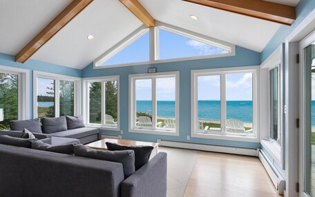 A spacious living room with a large gray sectional sofa, multiple windows offering ocean views, and a high ceiling with exposed wooden beams.