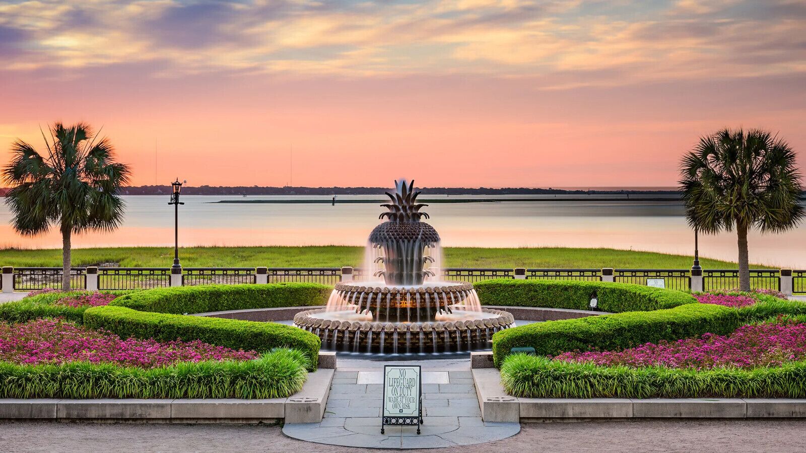 A fountain with water jets, surrounded by manicured hedges and flowering plants, with a view of a calm body of water and a sunset in the background.