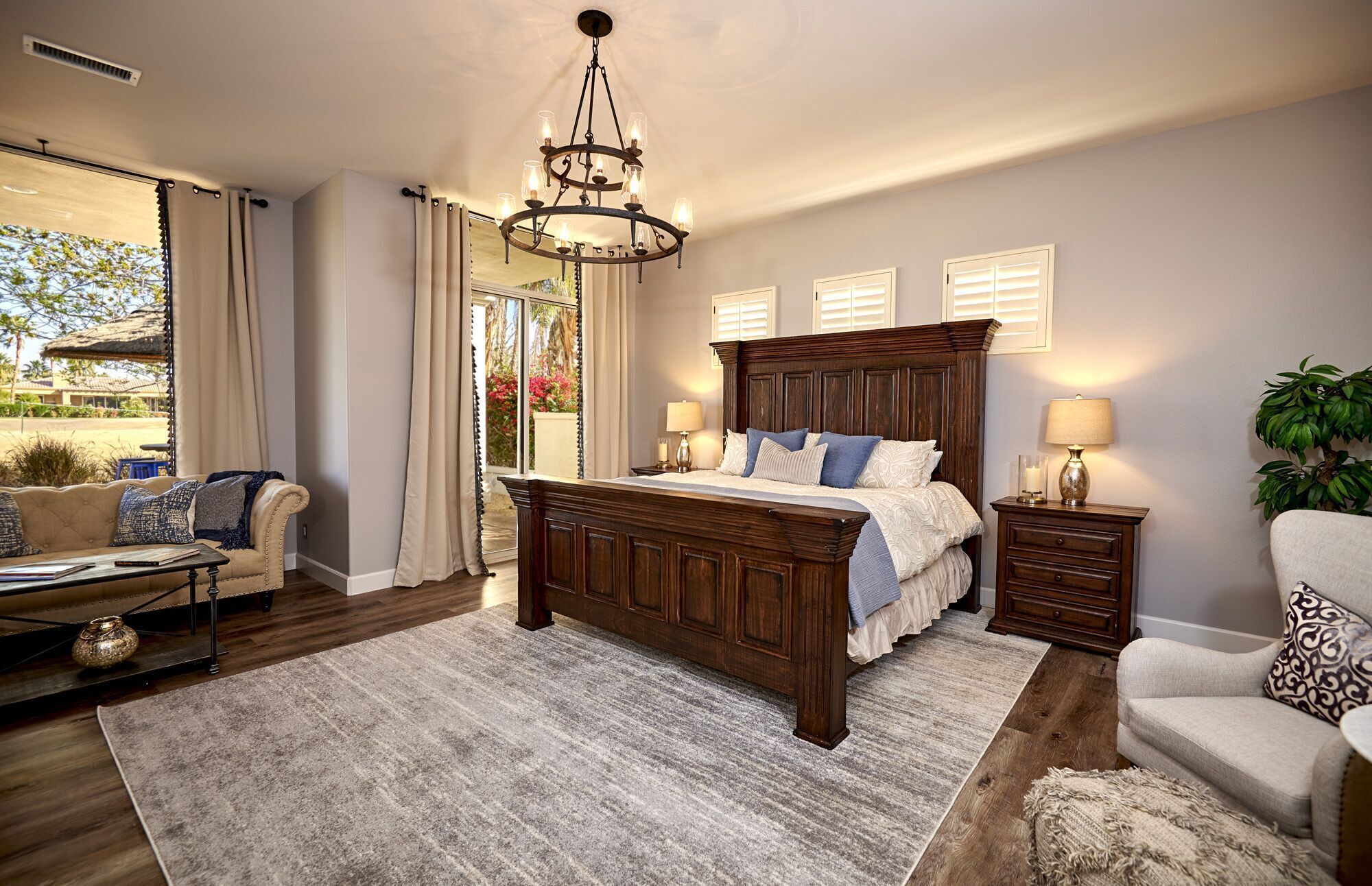 A bedroom with a large bed, a chandelier, a sofa, a chair, a nightstand, and a view of the outdoors.
