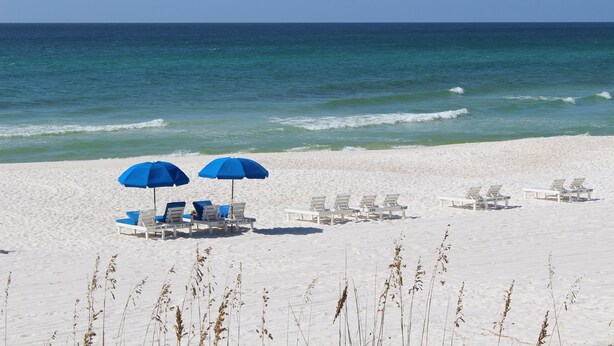 Een strand met wit zand en helderblauw water. Er staan meerdere witte ligbedden en twee blauwe parasols.