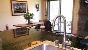 A kitchen with a stainless steel sink, a wooden table, a chair, a potted plant, and a framed picture on the wall.
