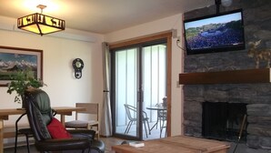 A living room with a fireplace, a television mounted on a stone wall, a leather chair, a wooden coffee table, and a dining area with a table and chairs.