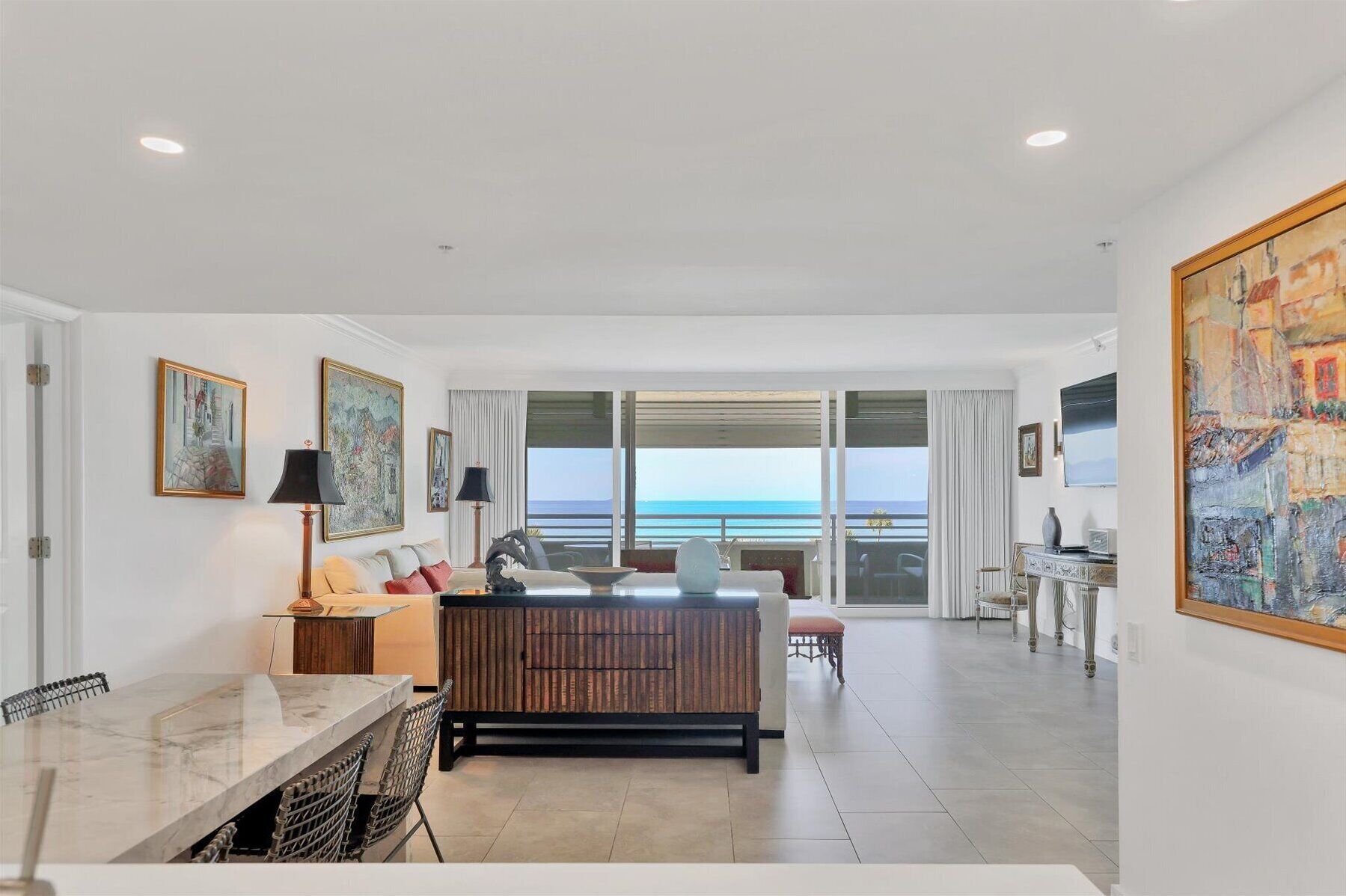 A spacious living area with a large window overlooking the sea, a dining area with a marble table, and a cozy seating area with a wooden bench.