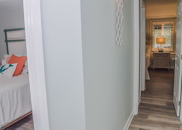 A hallway with a bed on the left and a lamp in the room at the end.