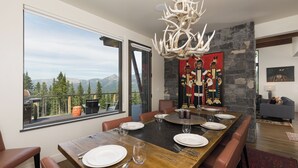 Un coin repas avec une grande table en bois dressée pour six personnes, un lustre et une vue sur les montagnes par de grandes fenêtres.