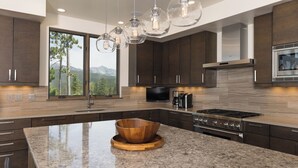 Une cuisine moderne avec des meubles en bois foncé, un grand îlot central agrémenté d’un saladier en bois, des appareils électroménagers en acier inoxydable et une vue sur les montagnes grâce à de grandes fenêtres.