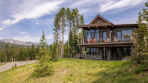 Une maison moderne à deux étages, dotée d’un grand balcon, entourée de grands pins et offrant une vue sur un paysage montagneux.
