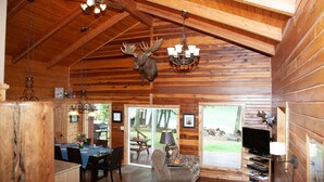 Interior rústico de una cabaña con comedor, chimenea, televisor y una decoración de cabeza de venado colgada del techo.