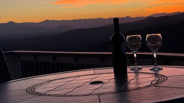 Une table avec une bouteille et deux verres sur un balcon donnant sur un paysage montagneux au coucher du soleil.