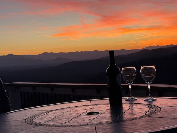 A table with a bottle and two glasses on a balcony overlooking a mountainous landscape during sunset.