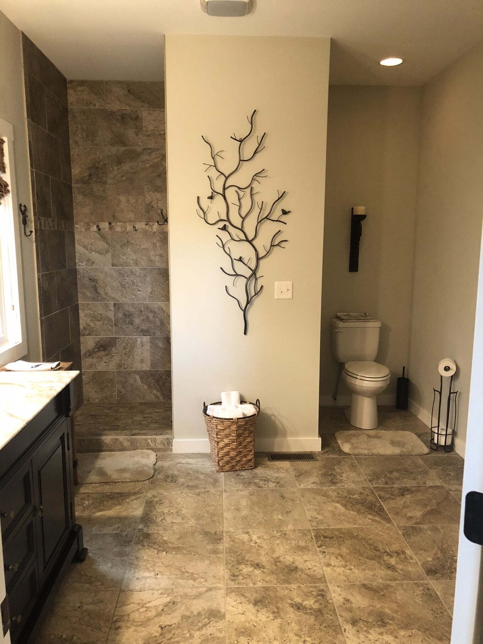 Une salle de bain avec un mur en pierre, une décoration murale représentant une branche d'arbre, un panier en osier, des toilettes et un lavabo avec un meuble sombre.