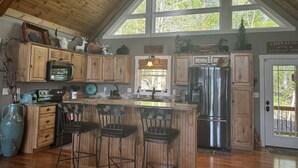 Une cuisine spacieuse avec des meubles en bois, un grand îlot central doté de tabourets, des appareils en acier inoxydable et une vue sur les arbres grâce à de grandes fenêtres.