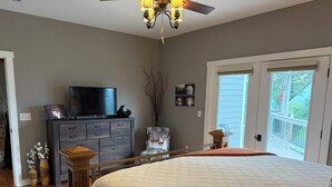 Une chambre avec un grand lit, une commode, une télévision et une chaise. Il y a un balcon avec une balustrade et une porte donnant sur l’extérieur.