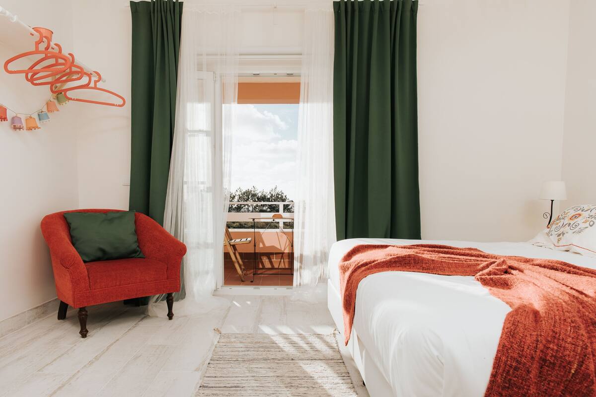 Una camera da letto con un letto, una poltrona rossa, un balcone con tavolo e sedie e tende verdi.