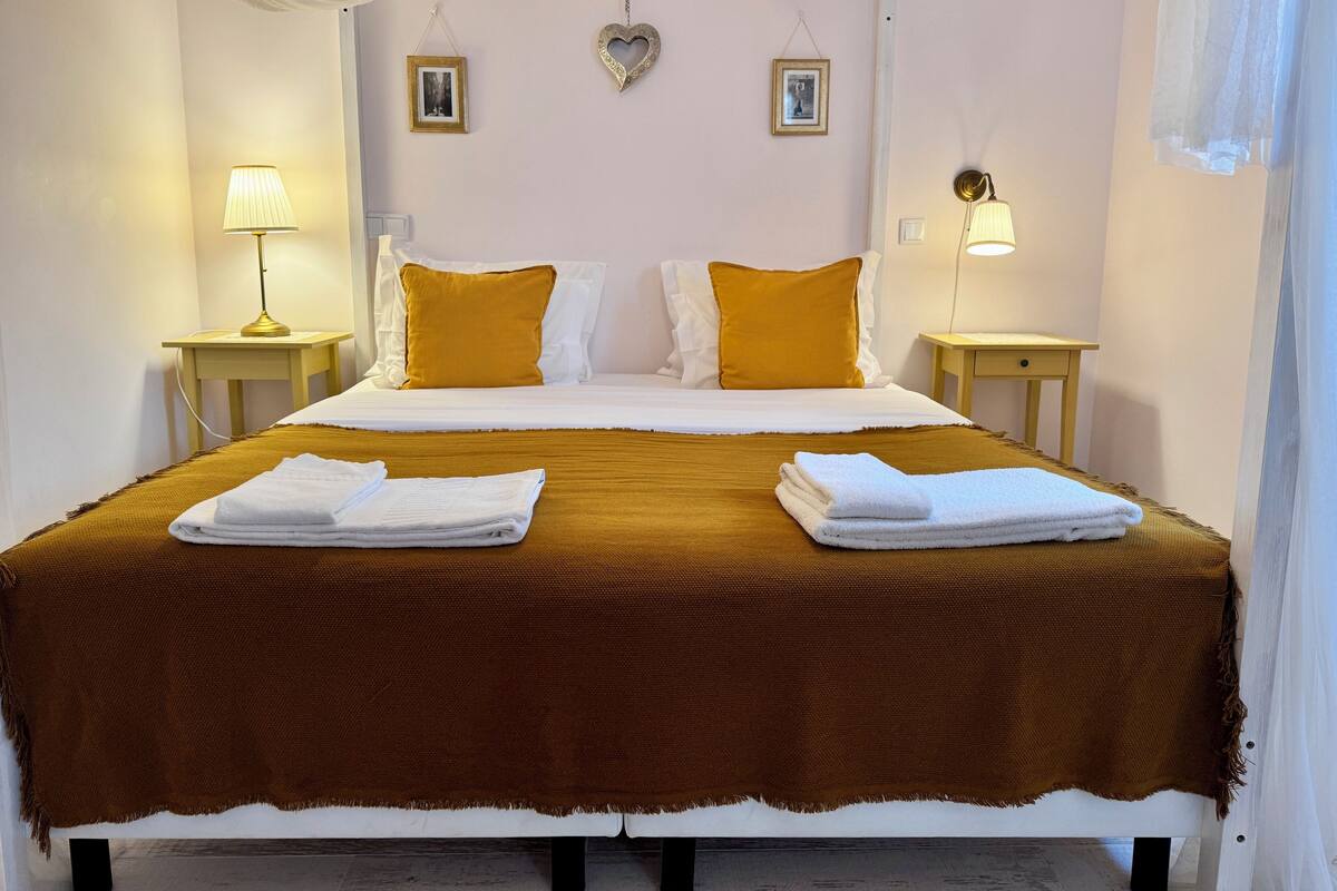 Un letto rifatto con cura, con una coperta marrone, cuscini bianchi e due asciugamani piegati. Su due comodini in legno ci sono due abat-jour e due decorazioni a forma di cuore appese al muro.