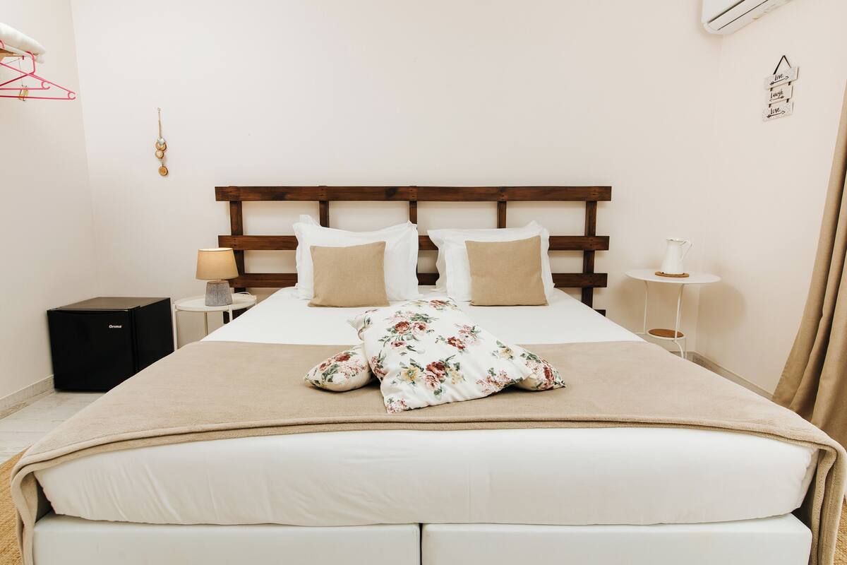 Un letto rifatto con cura, una coperta floreale e due cuscini, un comodino con una lampada e un piccolo elettrodomestico nero sul pavimento.