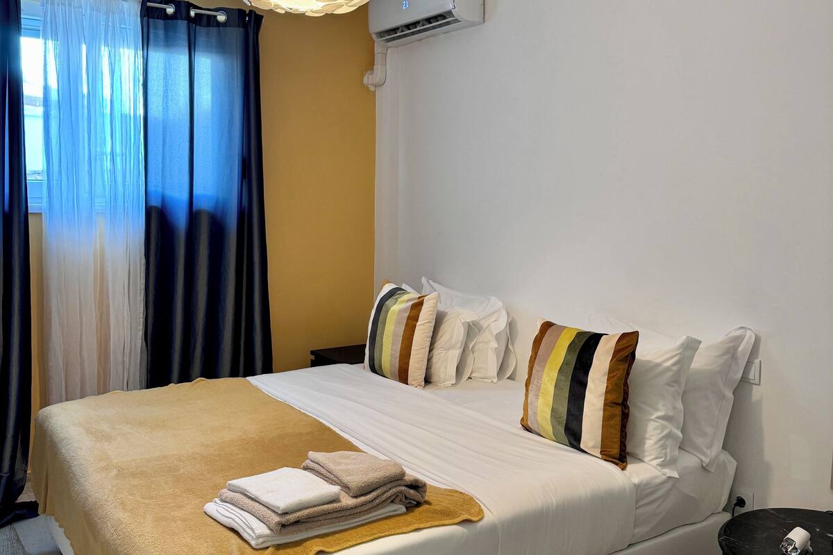 Un letto ben rifatto con coperta e cuscini color beige, un comodino con una lampada e un condizionatore d'aria a parete.