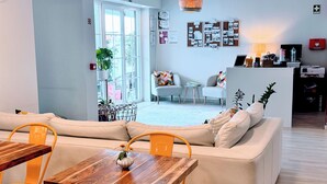 A modern living room with a sofa, wooden dining table, and yellow chairs. There is a kitchen area with a counter and appliances visible in the background.