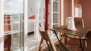 A balcony with a wooden table and chairs, a white pitcher, and a red curtain.