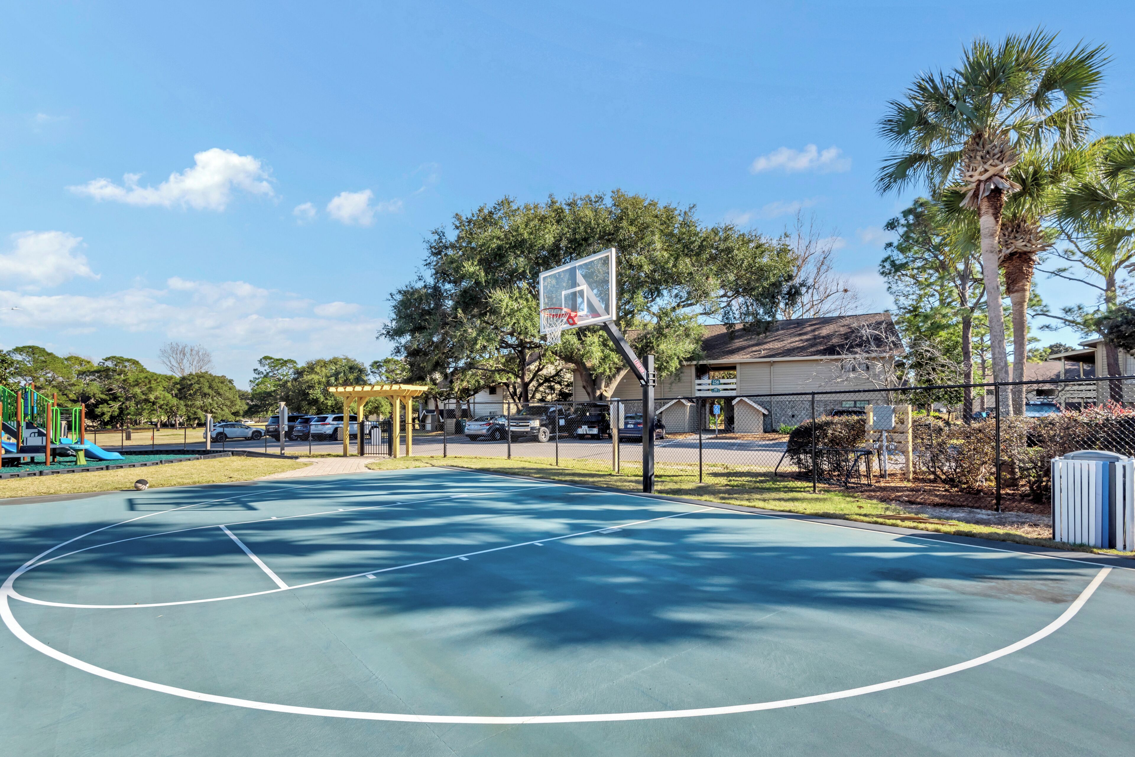 Ein Basketballplatz im Freien mit Korb, umgeben von Bäumen und einem Spielplatz.