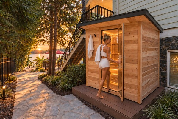 A wooden sauna structure with a glass door, a stone pathway, and a view of a body of water in the background.