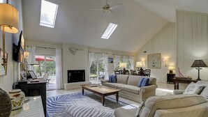 Amplia sala de estar con televisor de pantalla plana, un sofá, una mesa de centro y chimenea. Hay dos ventanas con cortinas, un ventilador de techo y un comedor con una mesa y sillas.