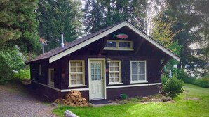 Une petite cabane à un seul étage, avec une porte et des fenêtres blanches, entourée d’une cour verdoyante et d’arbres.