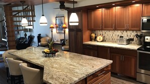 Una cocina moderna con encimeras de granito, armarios de madera y electrodomésticos de acero inoxidable. Hay luces colgantes, un frutero y una zona de comedor con taburetes de bar.