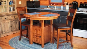 Une table de salle à manger en bois, accompagnée de deux chaises, un coin cuisine équipé d’appareils électroménagers et un meuble contenant divers objets.