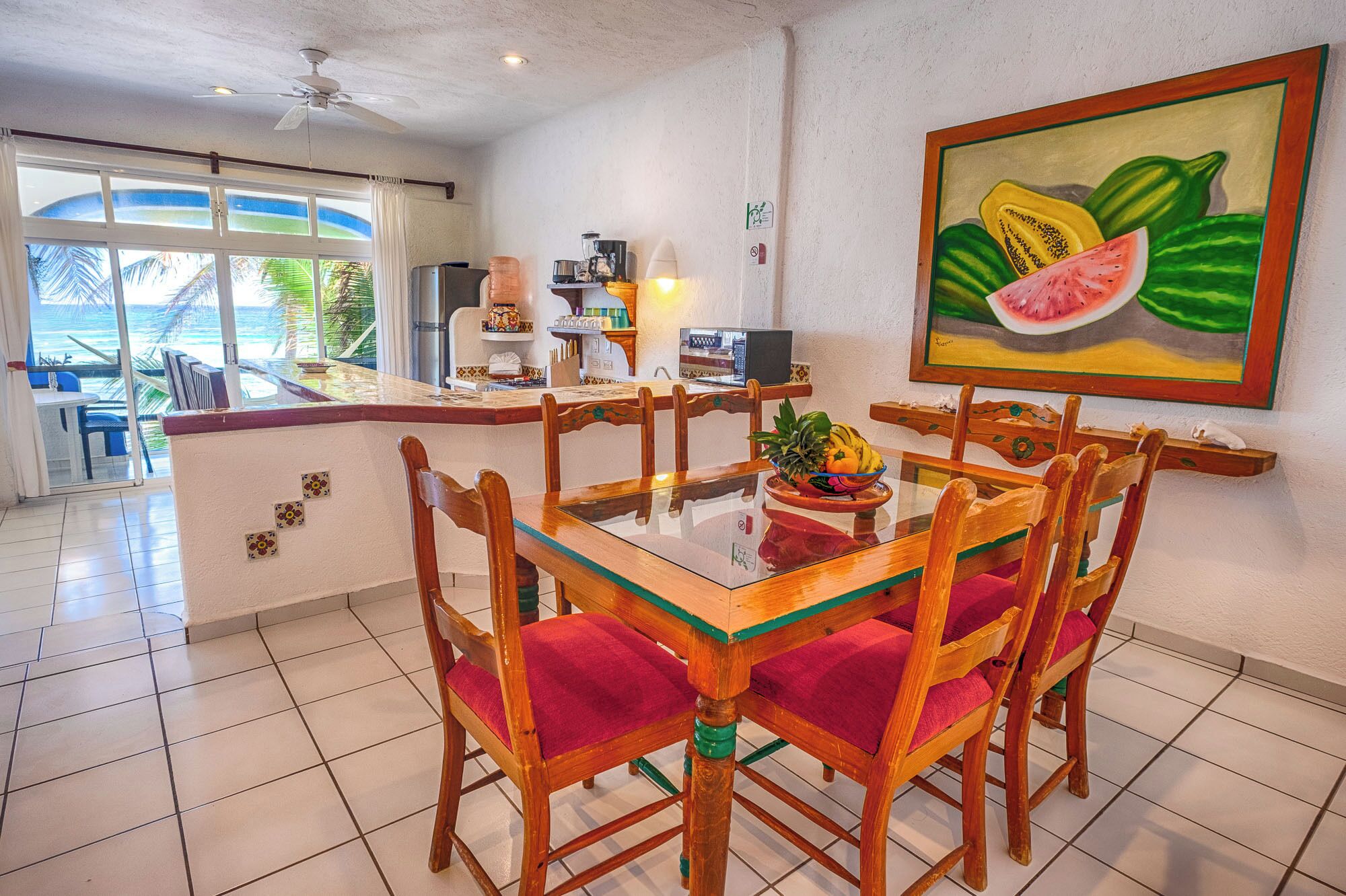A dining area with a glass table and wooden chairs, a painting of fruit on the wall, and a view of the ocean through large windows.
