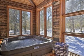 Une cabane en rondins dotée d’un bain à remous et de grandes fenêtres donnant sur une forêt enneigée.