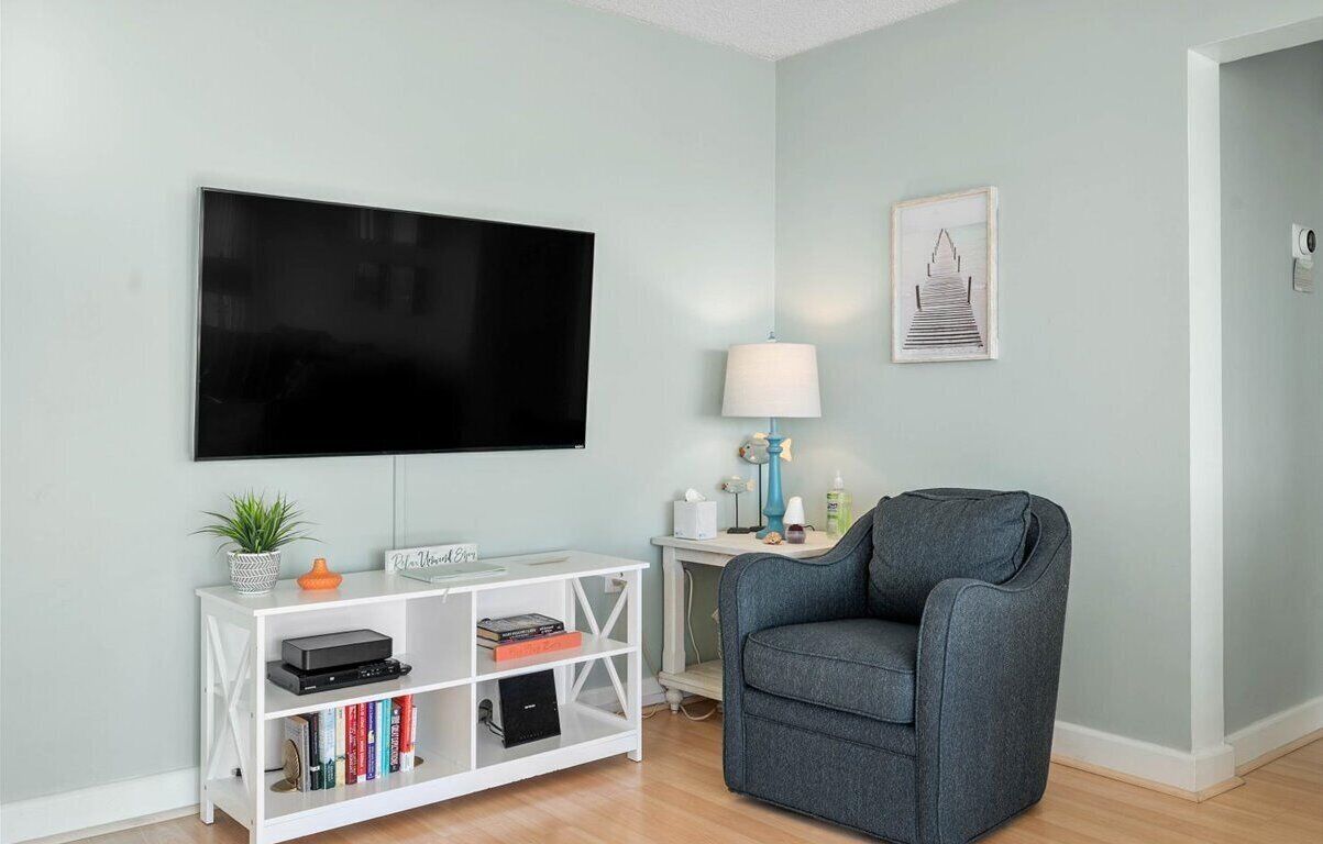 Ein Wohnzimmer mit Flachbildfernseher, einem grauen Sessel, einem weißen Konsolentisch mit Büchern und einer Pflanze sowie einem gerahmten Bild an der Wand.