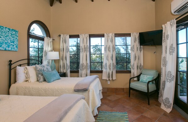 A bedroom with two beds, a chair, a TV, and large windows with curtains.