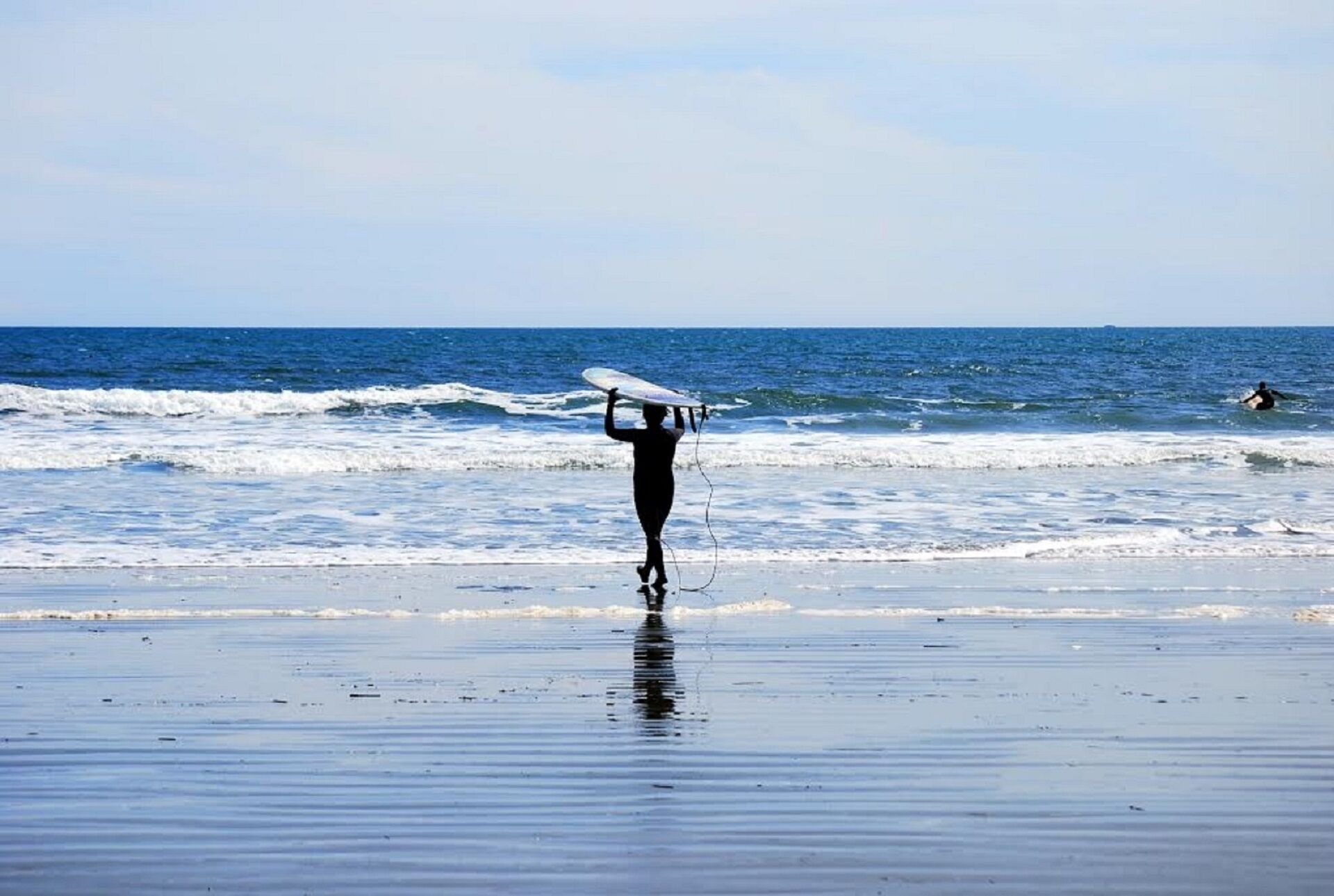 A beach with a person carrying a surfboard, waves, and another person surfing in the background.