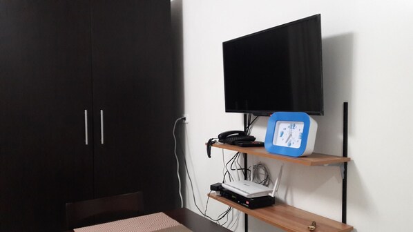 A room with a dark wardrobe, a desk with a chair, a mounted TV, a blue clock, and a shelf with electronic devices.