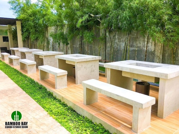 A series of concrete benches and tables arranged outdoors, with a bamboo wall in the background.