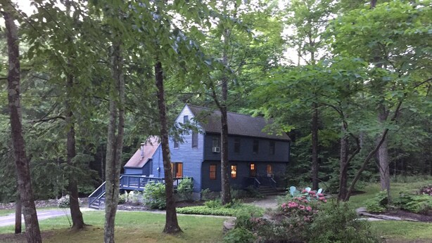 A two-story house with a dark blue exterior, surrounded by a forested area with a well-maintained garden and a small bridge leading to the house. - <br>Escape to our incredibly private home on 30 acres! (Saugerties)