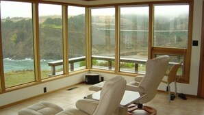 Ein geräumiges Zimmer mit großen Fenstern und Blick auf Meer und Hügel. Zwei Stühle und ein kleiner Tisch sind vorhanden.