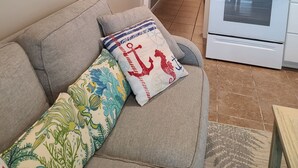 Ein Wohnzimmer mit grauem Sofa, einer Wanddekoration und einer Küchenzeile mit weißen Geräten.