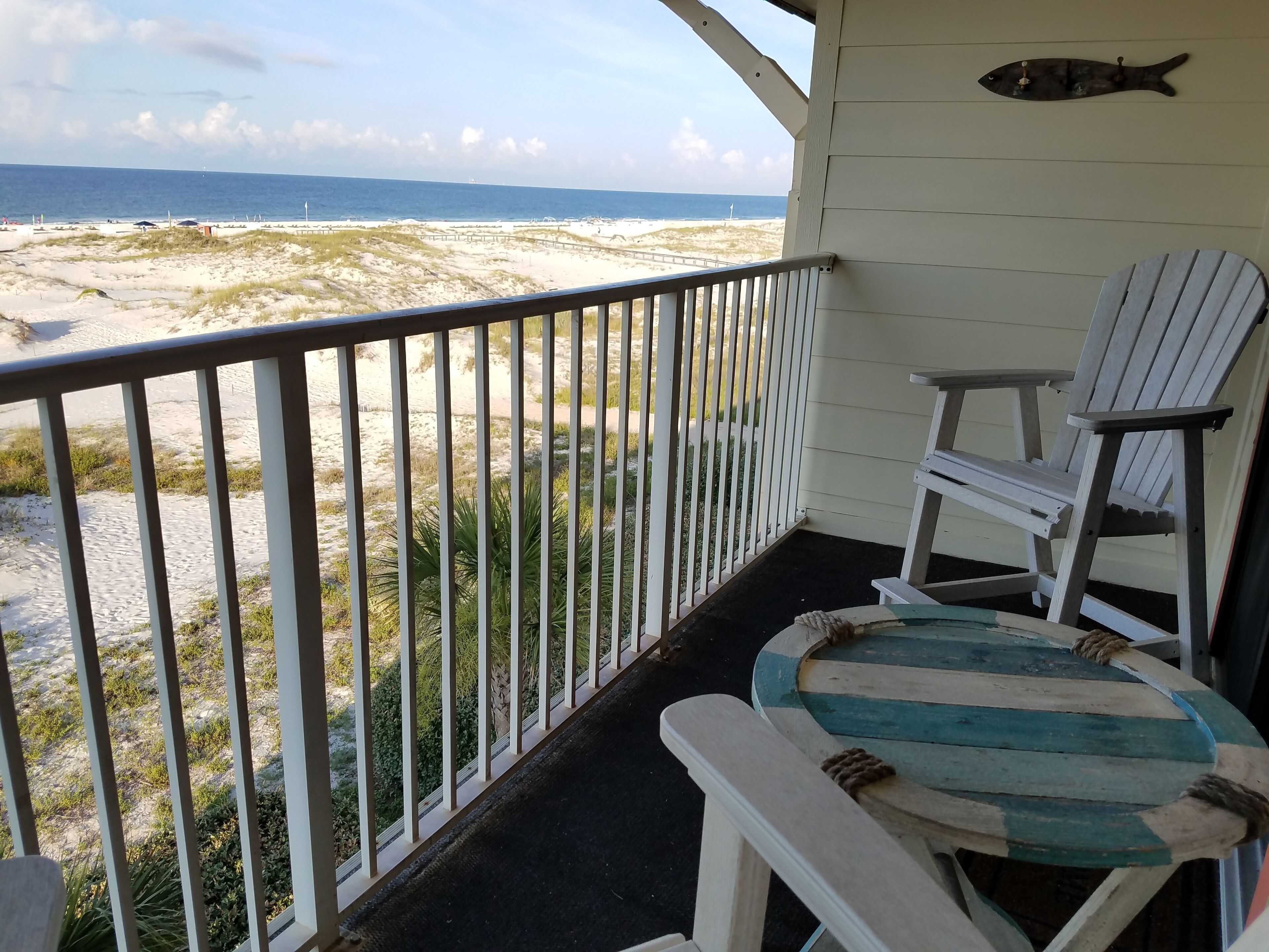 A balcony with a view of the beach, a fish decoration, and two Adirondack chairs.