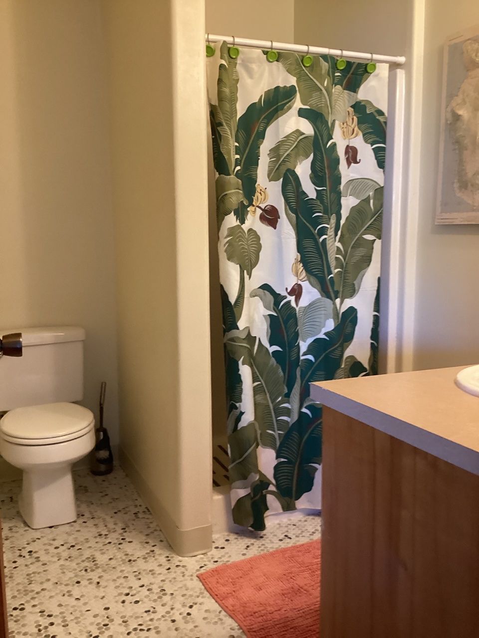 Un baño con cortina de ducha estampada con hojas tropicales, un inodoro blanco, un lavabo de madera y un suelo moteado.
