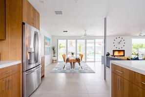 A modern kitchen with wooden cabinets, a stainless steel refrigerator, and a dining area with a table and chairs. - Burton Bliss Solar Powered Desert Home in Sunrise Park (Palm Springs)